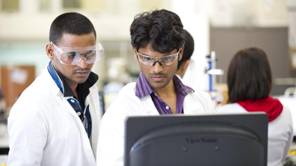 实验室外套和护目镜中的两名年轻男性科学家站着看计算机监视器。