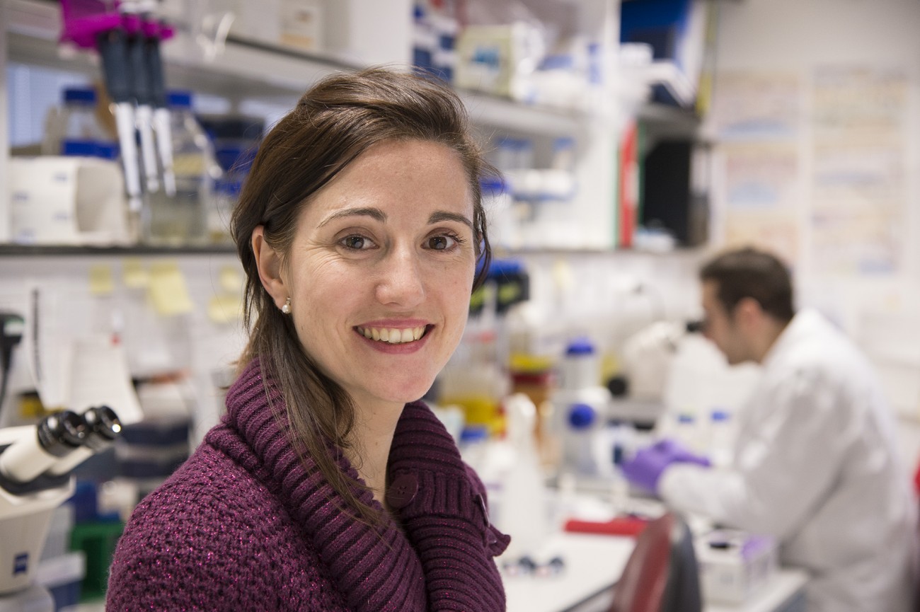 Meritxell uch博士，2013年3Rs奖得主。她在一个实验室里，你可以看到一个穿着白色外套和紫色手套的研究人员在背景中俯视着显微镜。
