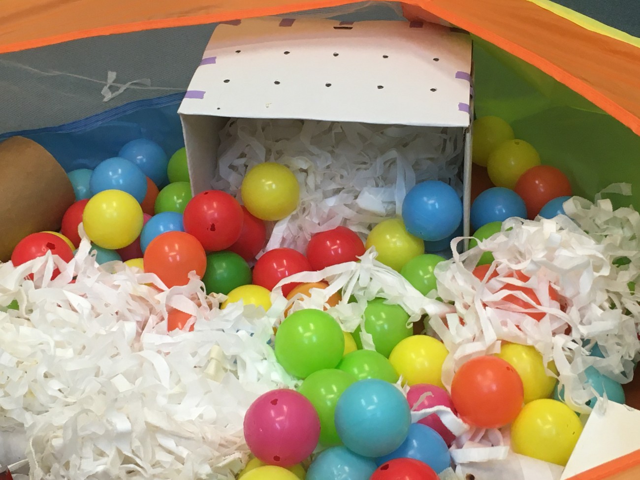 一个穿孔的纸板箱放在游戏围栏里。载体里装满了条状的垫层材料，周围环绕着色彩鲜艳的球。
